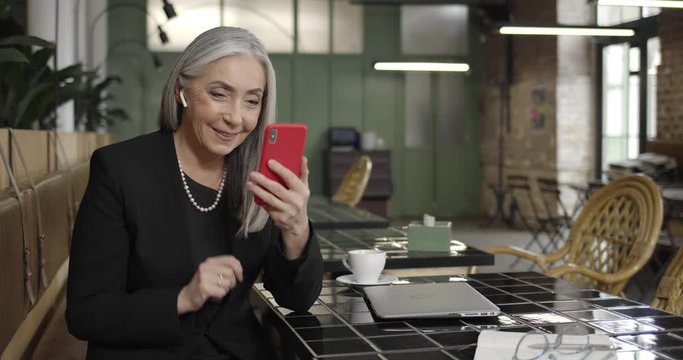 Front view of mature lady enterpreneur in earpods having videocall on smartphone with business partner .Middle aged woman communicating, using video app while sitting in cafe .
