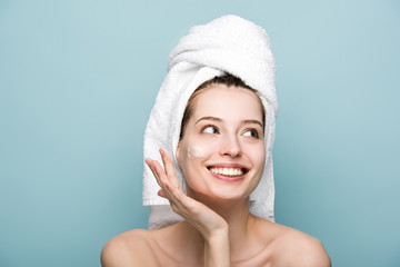 happy attractive girl applying moisturizing facial mask while looking away isolated on blue