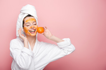 happy girl with citrus facial mask holding tangerine near face on pink background