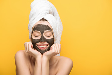 cheerful girl with facial clay mask and towel on head holding hands near face with closed eyes isolated on yellow