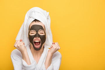 irritated girl with facial clay mask and towel on head holding clenched fists and screaming isolated on yellow