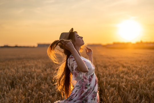Incredible young woman with long curly hair. Woman in dress posing in wheat field at sunset and straightens hair tilting head back
