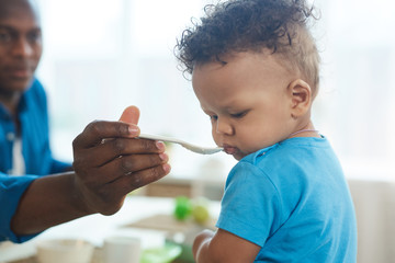 Side view portrait of cute African-American toddler refusing to eat food from spoon in kitchen interior, copy space