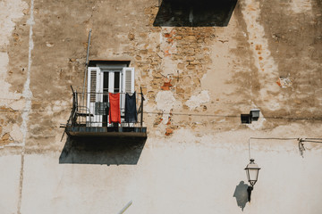 Red and blue dresses hanging on the metal railing of the small balcony of the old house with the wall with cracked facade in Slovenia coastal town Piran during the sunny summer day