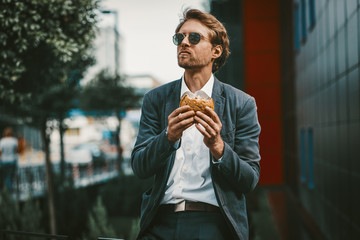 Handsome trainee having a lunch break near his office. Young man eats hamburger outside of his business center. Non healthy food concept. - Powered by Adobe