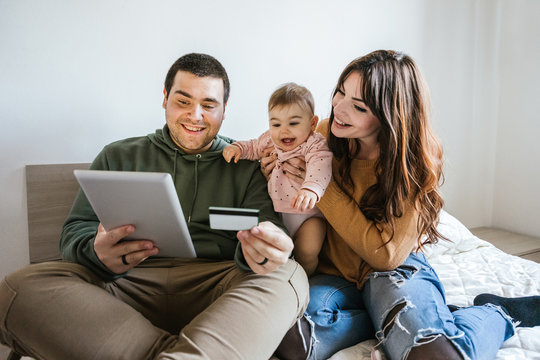 Portrait of a family on the bed looking at the tablet with their baby girl and shopping online with a credit card - Father, mother and little daughter have fun together - Intimacy moment