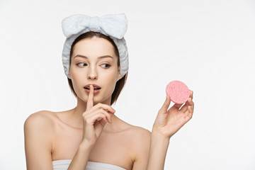 attractive girl showing hush gesture while holding cosmetic sponge isolated on white