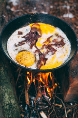Fried eggs with bacon in a pan in the forest. Food at the camp. Fried egg with bacon on fire. Picnic