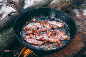 Slices of fried bacon in a pan. Food in a forest camp. Cooking on fire. Picnic in the nature. Grilled food on nature..