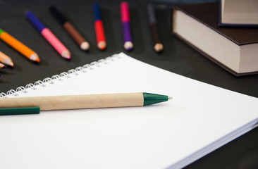 Education concept. Notebook with pen on the background of colored pencils and books on a black table. Close-up.