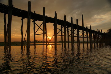 Beautiful sunset view of teakwood U Bein Bridge on background of bright orange sky, reflection in water of lake, Burma
