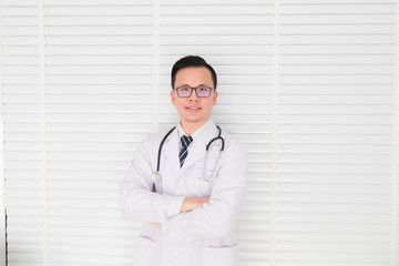 Thai doctor, an Asian man, wears glasses Hanging the stethoscope and wearing a uniform, smiling and standing, folded across the hospital examination room