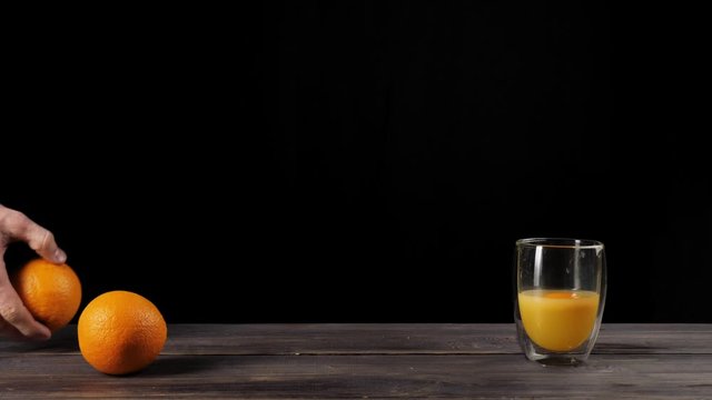 Wide shot of hand takes oranges from the table and throws them in a double-walled glass in turn, after the oranges reach the glass, orange juice appears against black background