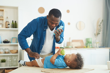 Portrait of mature African-American man calling wife while changing diaper to baby in home interior, copy space - obrazy, fototapety, plakaty