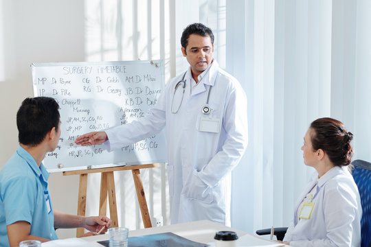 Serious mature Indian general practitioner showing surgery timetable on whitebord to colleagues