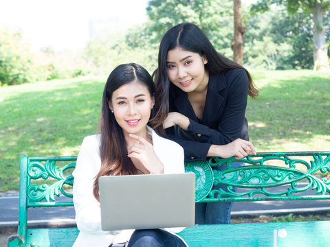 two asia business woman working with computer laptop in park. beauty woman tpying keyboard notebook blank screen and friend standing behind with copy space.  
