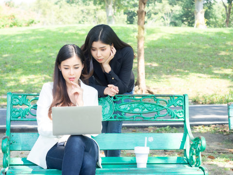two asia business woman working with computer laptop in park. beauty woman tpying keyboard notebook blank screen and friend standing behind with copy space.  