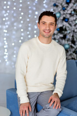 Handsome man wearing white sweater sitting on the lue sofa. Comfort and relaxation on gray background with bokeh