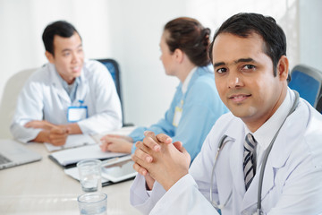 Fototapeta premium Portrait of smiling mature Indian doctor sitting at big table with colleagues discussing epidemic situation
