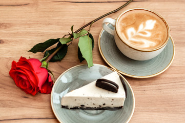 a piece of sweet dessert cake, red rose flower and a cup of cuppuccino on a wooden table. Romantic breakfast dinner, birthday, Valentine's Day, wedding, lovers, couple, romance.  Soft focus