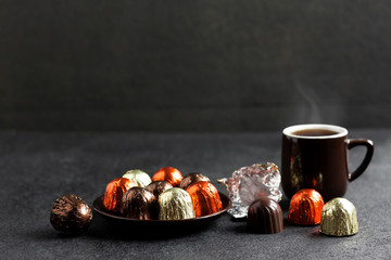 Chocolate candies wrapped in multicolored foil and cup of coffee on black background