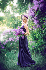 Outdoor fashion photo of a beautiful young woman surrounded by purple flowers. Girls in a long dress with a slit on the background of a spring garden with lilacs. The concept of cosmetics and perfumes