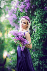 Outdoor fashion photo of a beautiful young woman surrounded by purple flowers. Girls in a long dress with a slit on the background of a spring garden with lilacs. The concept of cosmetics and perfumes