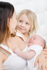 Young mother holding a newborn son in her arms smiling at her elder daughter with white hair sitting next to her