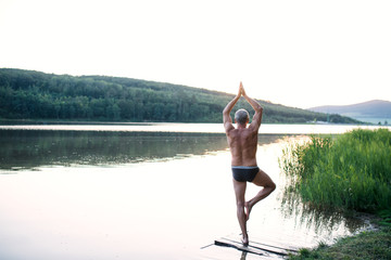 Rear view of senior man in swimsuit standing by lake outdoors doing yoga.