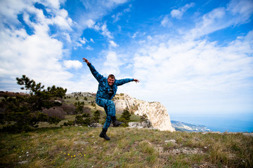 Funny young cheerful caucasian man jumping on the hills with green grass against a blue sky and white clouds. Concept of long-awaited travel and tourism