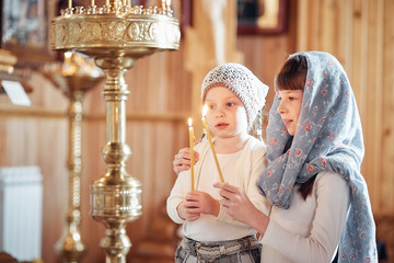 two Russian girls in a scarf on their heads stand in an Orthodox Church, lights a candle and prays in front of the icon.