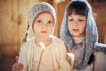 two Russian girls in a scarf on their heads stand in an Orthodox Church, lights a candle and prays...