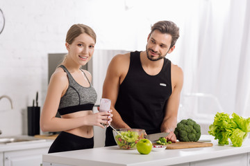 cheerful woman holding glass of smoothie near man cooking in kitchen