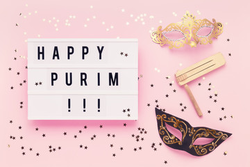 Flat lay of Purim Carnival celebration concept. Happy Purim written in light box, carnival mask and wooden gragger on pink background.