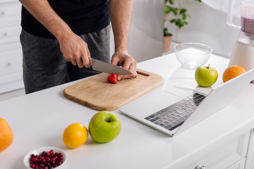 cropped view of man cutting strawberry near laptop