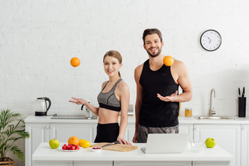 sportive girl and cheerful man throwing in air oranges near laptop