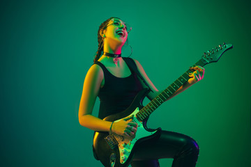 Rockstar. Caucasian female guitarist portrait on green studio background in neon light. Beautiful female model in black wear with guitar. Concept of human emotions, facial expression, ad, music, art.