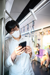Asian man wearing surgical face mask using smartphone on skytrain or urban train. Wuhan coronavirus (COVID-19) outbreak prevention in public transportation. Health awareness for pandemic protection