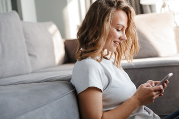 Image of happy woman smiling and using cellphone while sitting on floor