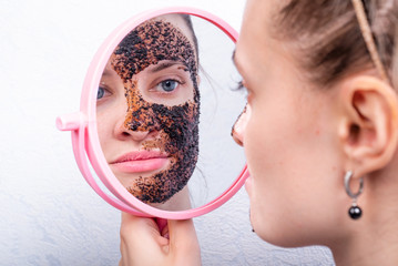 Young attractive woman is applying scrub on her face, looking in the mirror - coffee cleansing scrub