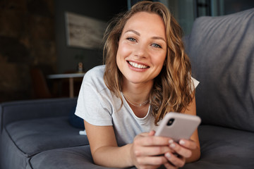 Image of happy woman smiling and using cellphone while lying on sofa