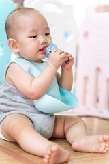 An Asian baby sits in a pen, biting a pacifier