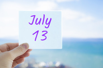 July 13th. Hand holding sticker with text July 13 on the blurred background of the sea and sky. Copy space for text. Month in calendar concept