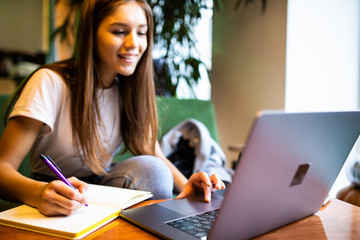 Young woman sitting at table and taking notes in notebook with table is laptop, smartphone and cup of coffee.