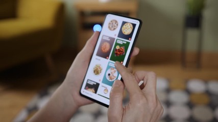 Orders Pizza Using Online Delivery. Woman Orders Food Home In Online Store Using a Smartphone. Female Ordering pizza using food delivery app. Screen is blurred. Focus on hand. - Powered by Adobe
