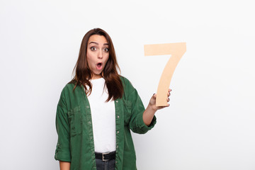 young pretty woman surprised, shocked, amazed, holding a number 7.