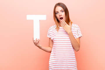 young pretty red head woman surprised, shocked, amazed, holding the letter T of the alphabet to...