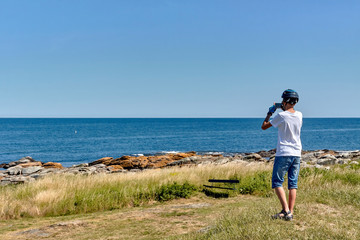 Rear view of a teenage boy wearing cycling helmet taking photos of the Baltic Sea with his smartphone, Svaneke, Bornholm Island, Denmark.