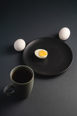 Boiled half egg with yolk in black plate near to white eggs and cup of tea on dark moody plain minimal background, angle view, happy Easter day