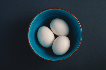White eggs in blue bowl on dark moody black plain minimal background, top view, happy Easter day